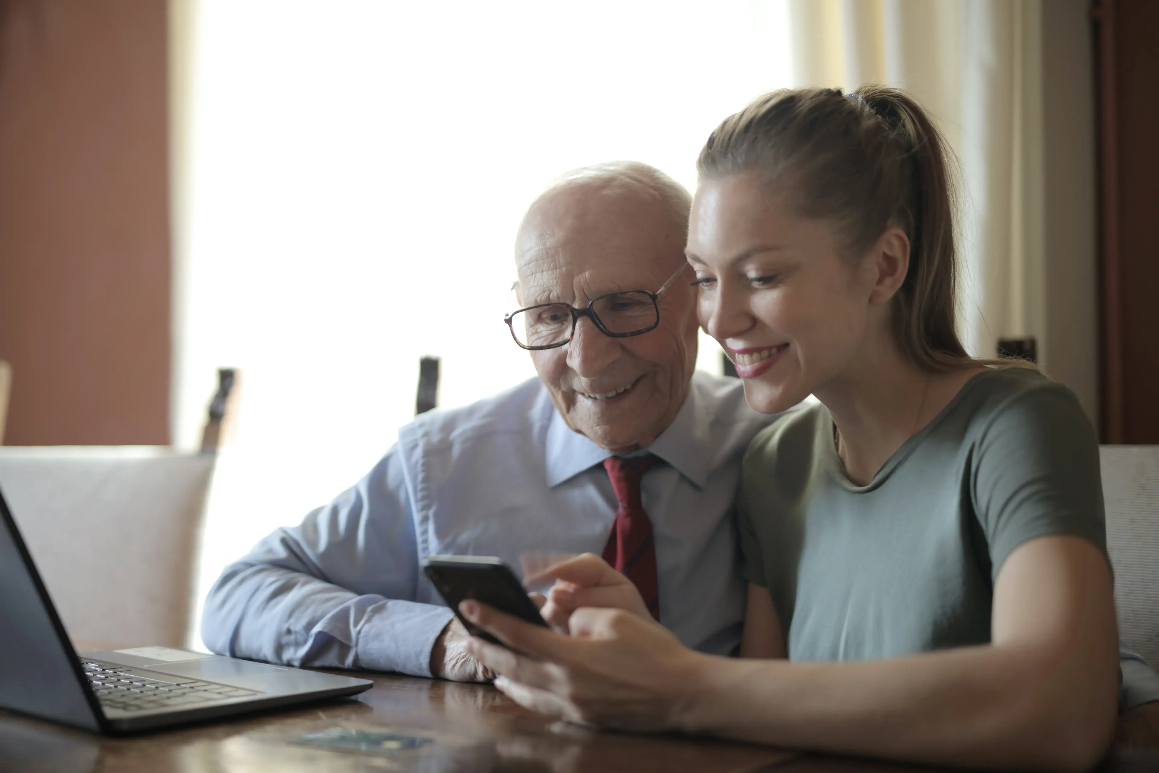 Smiling young woman in casual clothes showing smartphone to interested senior grandfather in a formal shirt and eyeglasses while sitting at a table near laptop reviewing health history